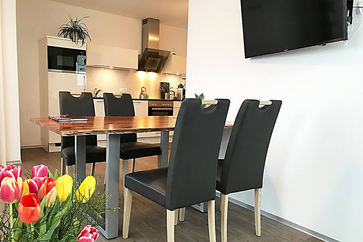 Een moderne eethoek met een houten tafel en zwarte stoelen, een keuken op de achtergrond en een flatscreen aan de muur.