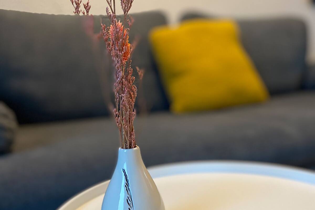 Een witte vaas met gedroogde planten op een witte tafel, voor een grijze bank met een geel kussen.
