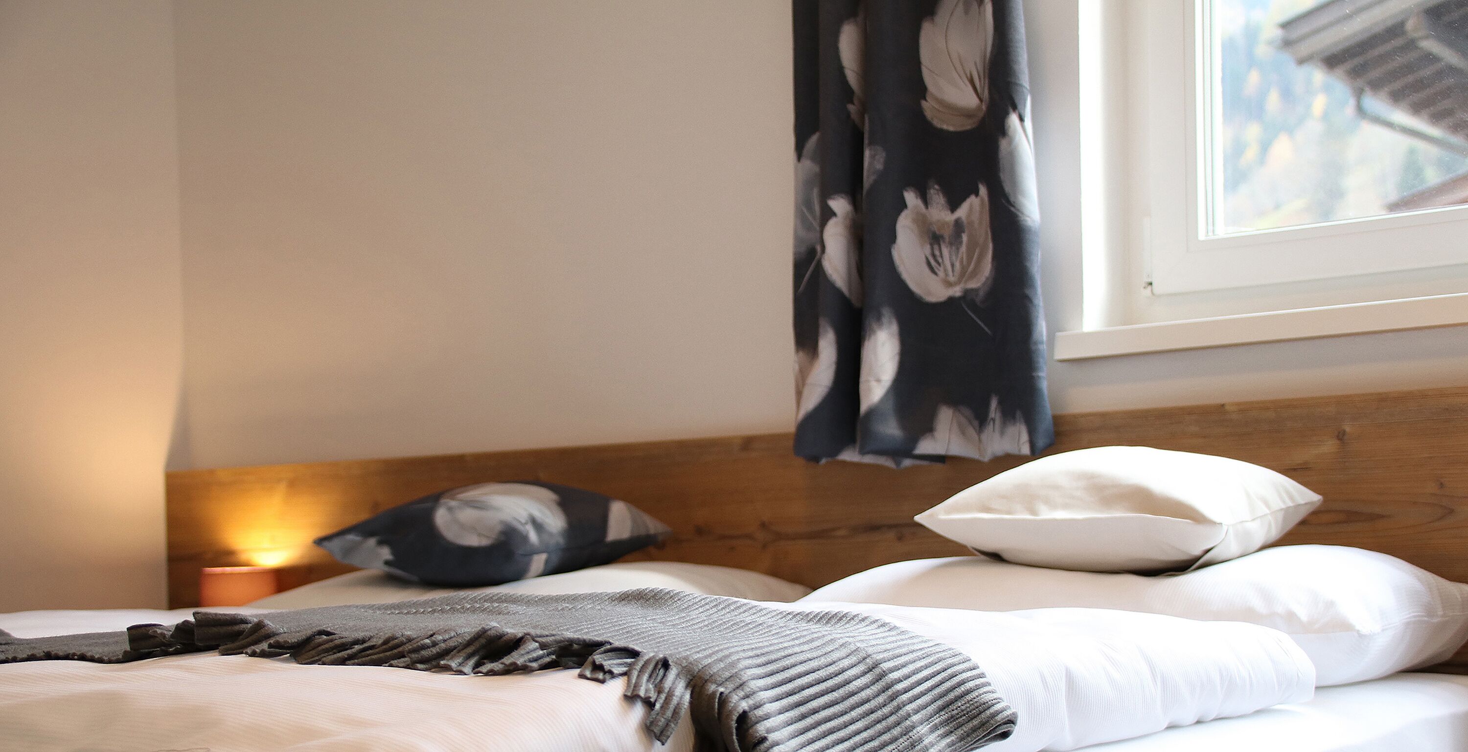 Una cama bien hecha con ropa de cama blanca y gris, un cabecero de madera y una ventana con vistas a edificios.