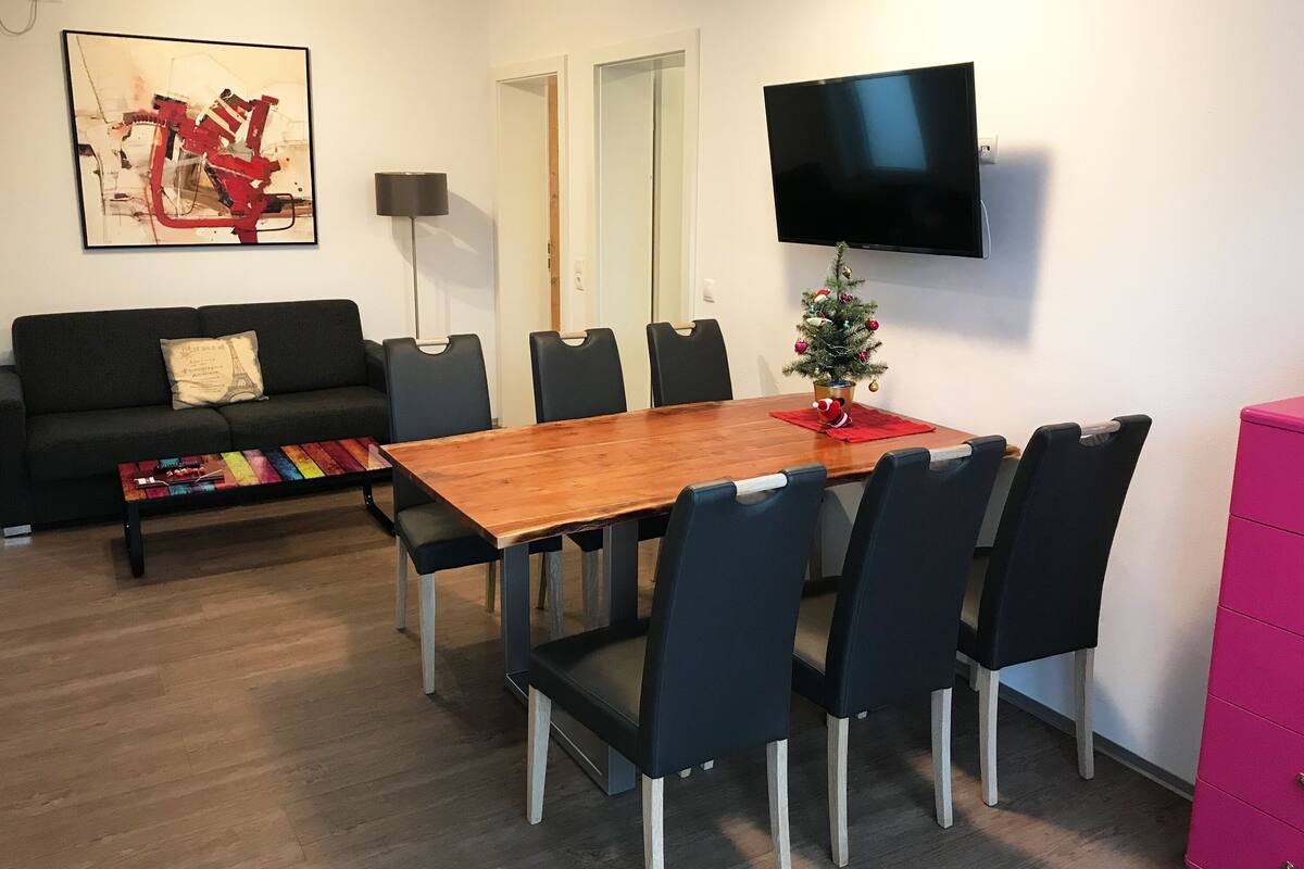 Een moderne woonkamer met een houten eettafel, zwarte stoelen, een zwarte bank, een kleurrijke salontafel, een kleine kerstboom en een flatscreen televisie aan de muur.
