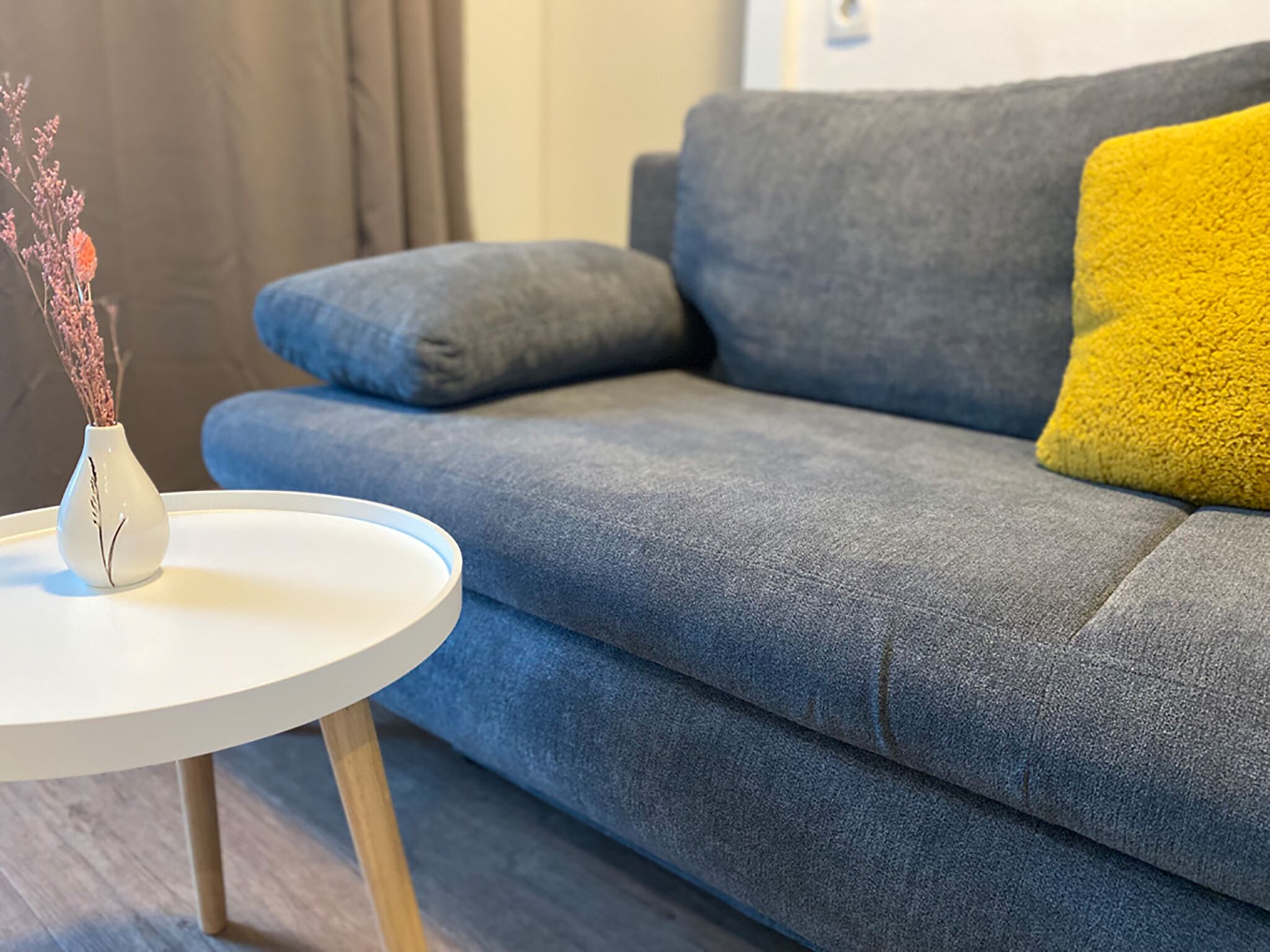 A modern living room with a grey sofa, a yellow pillow, and a white vase with dried flowers on a small round table.