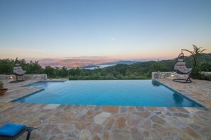 A infinity pool with stone flooring and two swing chairs, overlooking a mountainous landscape. - Villa Venetia (Corfu)