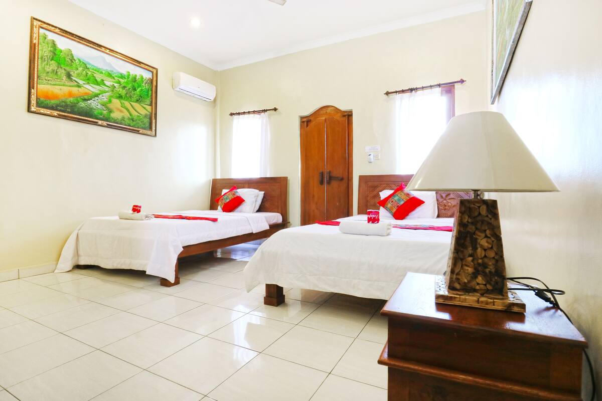 A hotel room with two beds, a wooden wardrobe, a painting on the wall, and a lamp on a wooden table.