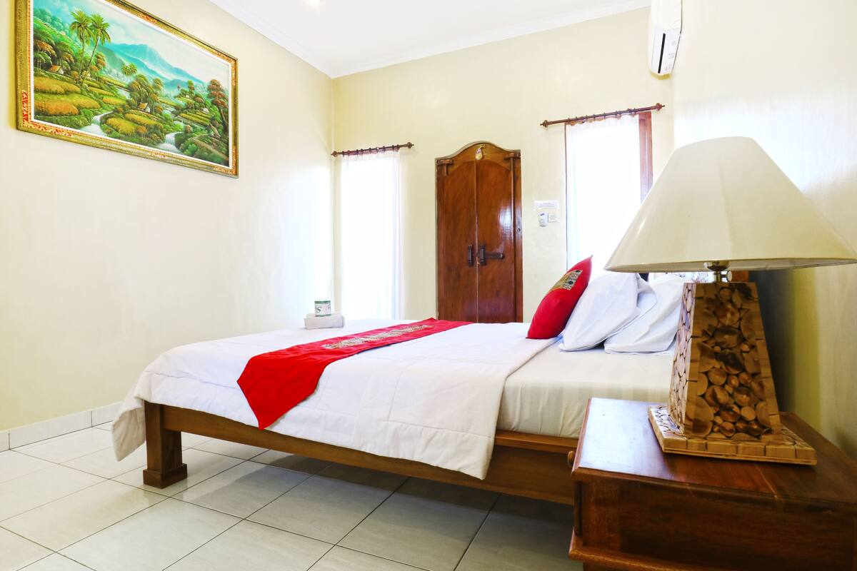 A neatly made bed with a red throw, a wooden nightstand with a lamp, a wooden wardrobe, and a painting on the wall.