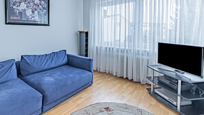 Een woonkamer met een blauwe bank, een flatscreen televisie op een standaard, een lijst met een foto aan de muur en dunne gordijnen voor de ramen.