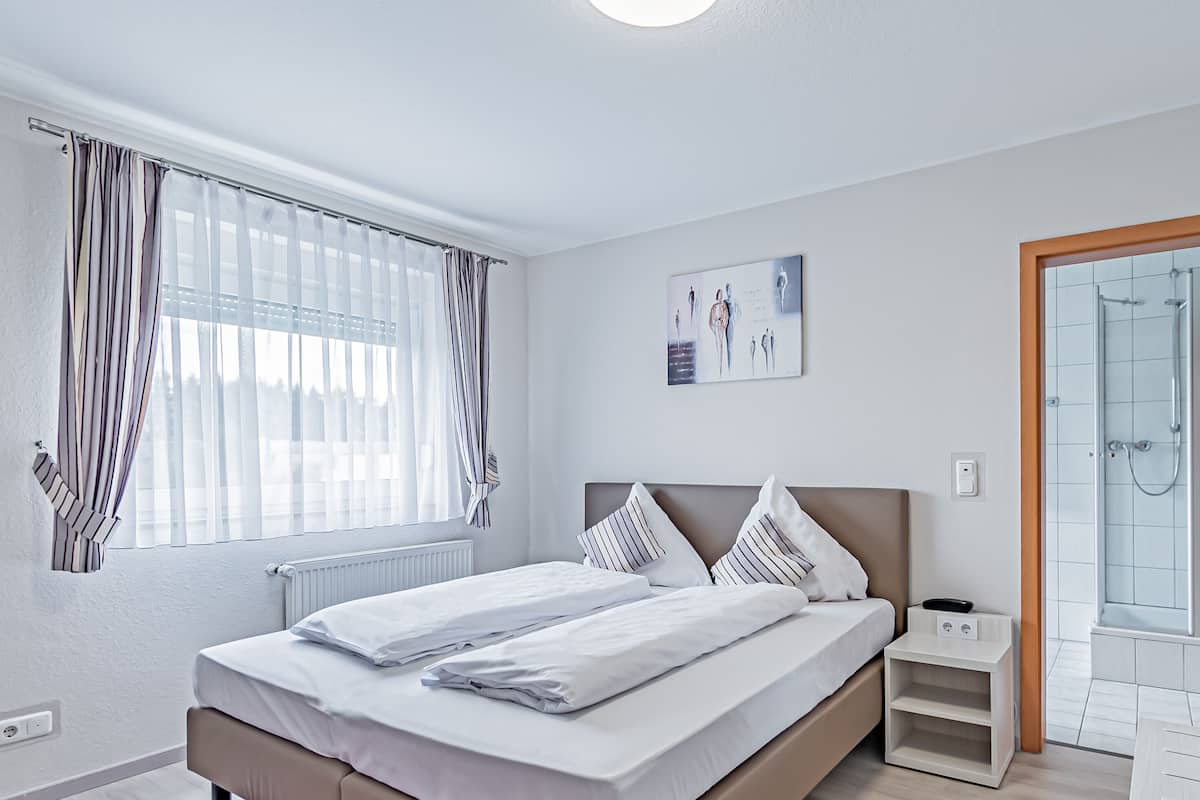 Quarto moderno com uma cama grande, mesa de cabeceira, área de chuveiro e janela com cortinas.