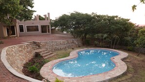 A swimming pool with a stone border, surrounded by a brick patio and a grassy area with trees. - Shiloh Luxury Lodge (Harare)