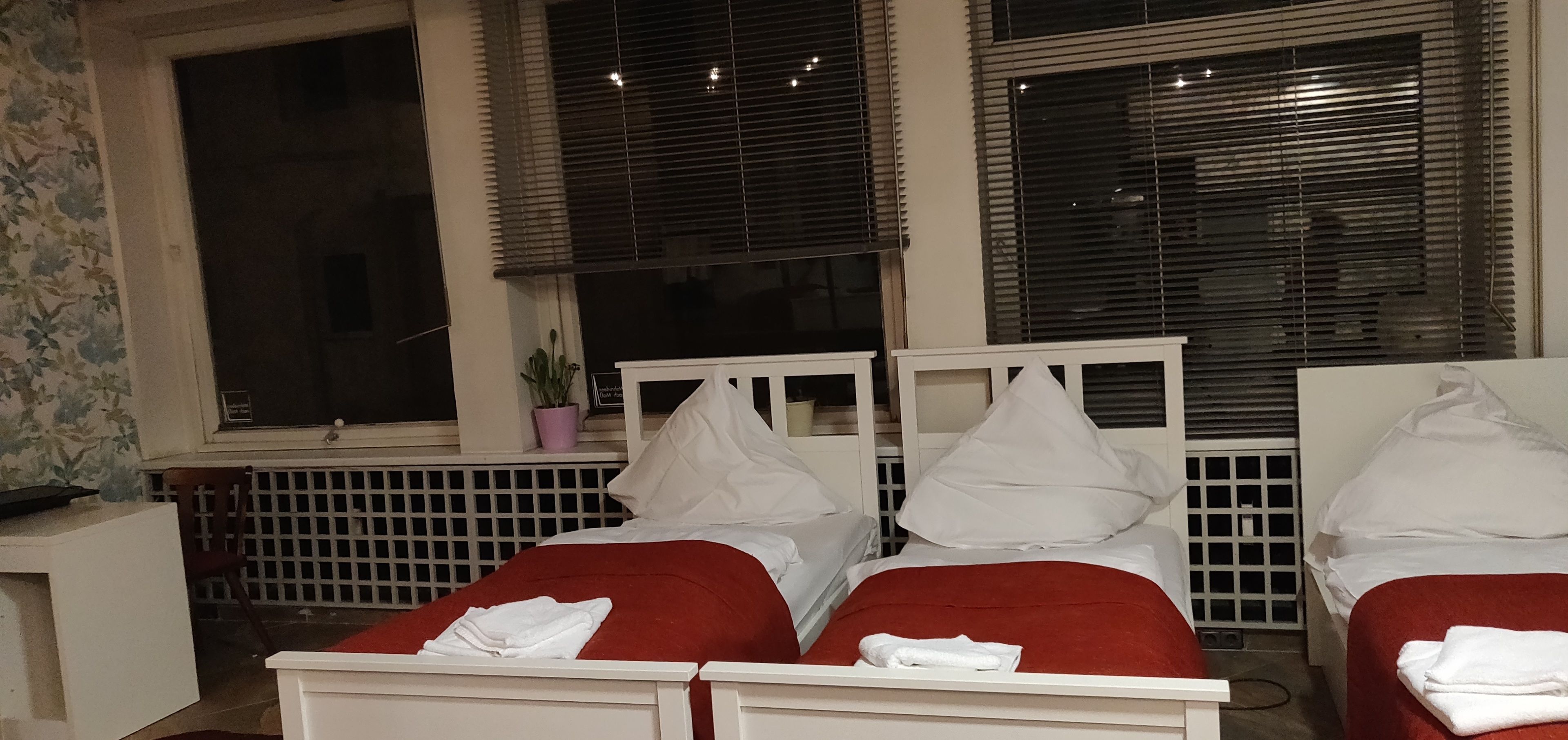 Habitación de hotel con dos camas, cada una con cabeceros blancos y ropa de cama roja. Se ve una ventana con persianas, una mesita con una planta en maceta y un radiador.