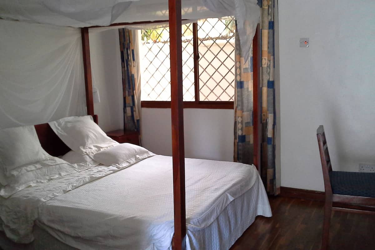 A single bed with a canopy, a wooden chair, and a window with a metal grill.