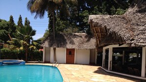 A swimming pool with a stone deck, a thatched-roof building, and palm trees. - Panori Hotel (Tanga)