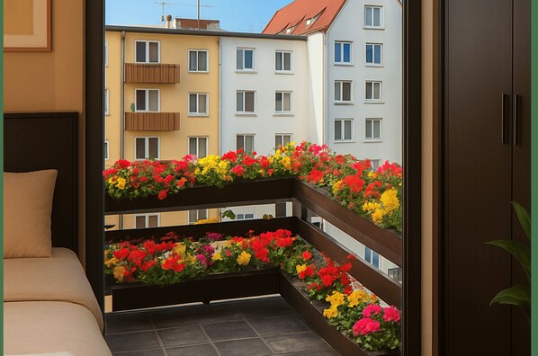 Un balcón con macetas de flores, vista a edificios de varios pisos y una puerta que da a una habitación.
