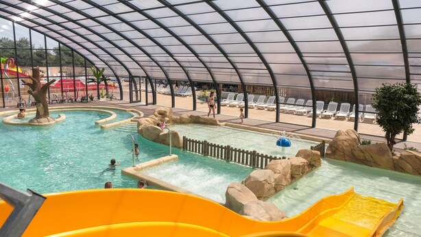 Parque acuático interior con múltiples piscinas, toboganes y una zona cubierta con sillones de descanso.
