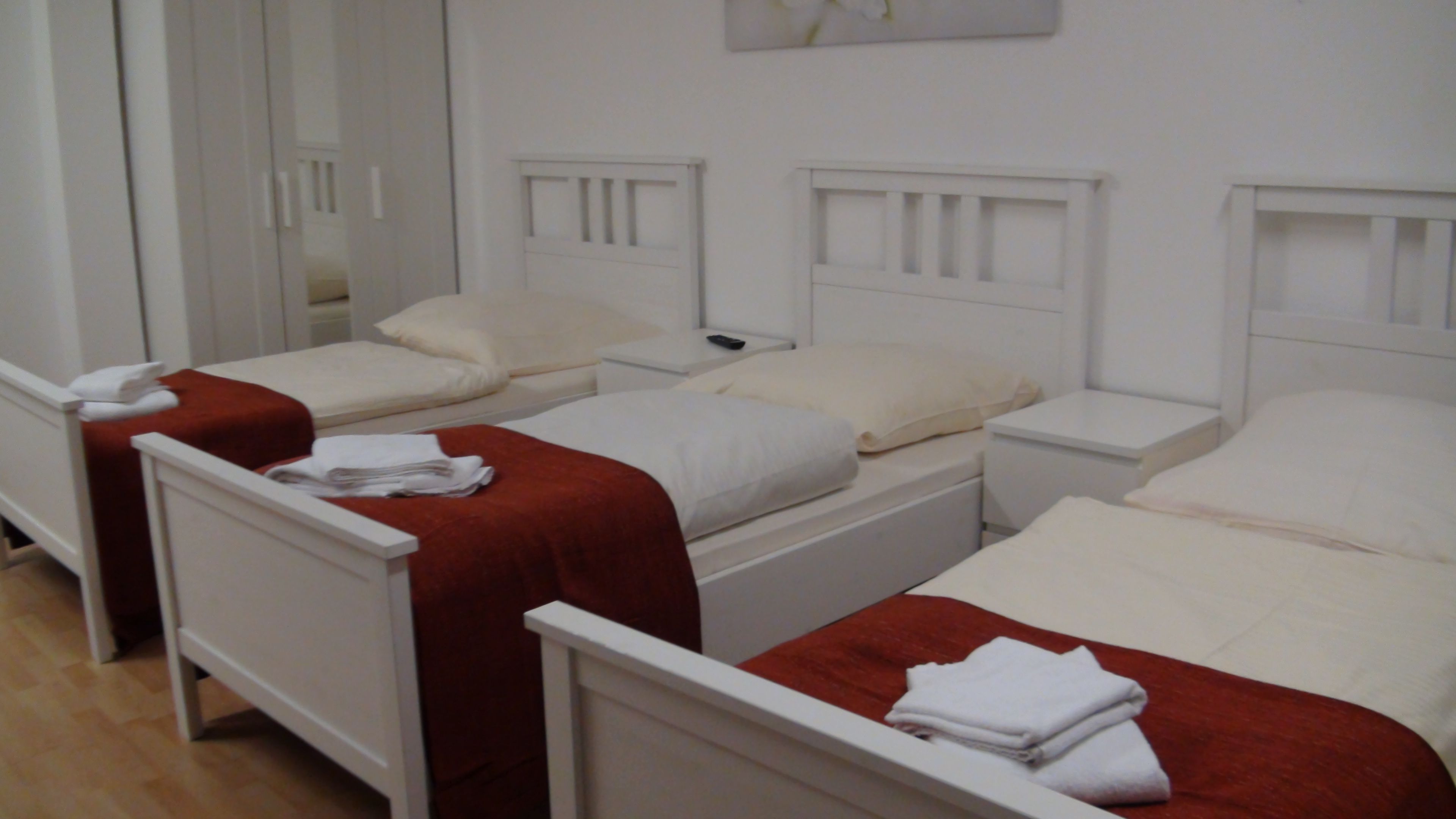 A hotel room with three single beds, each with a white headboard and a red blanket. There is a white nightstand with a remote control on it.