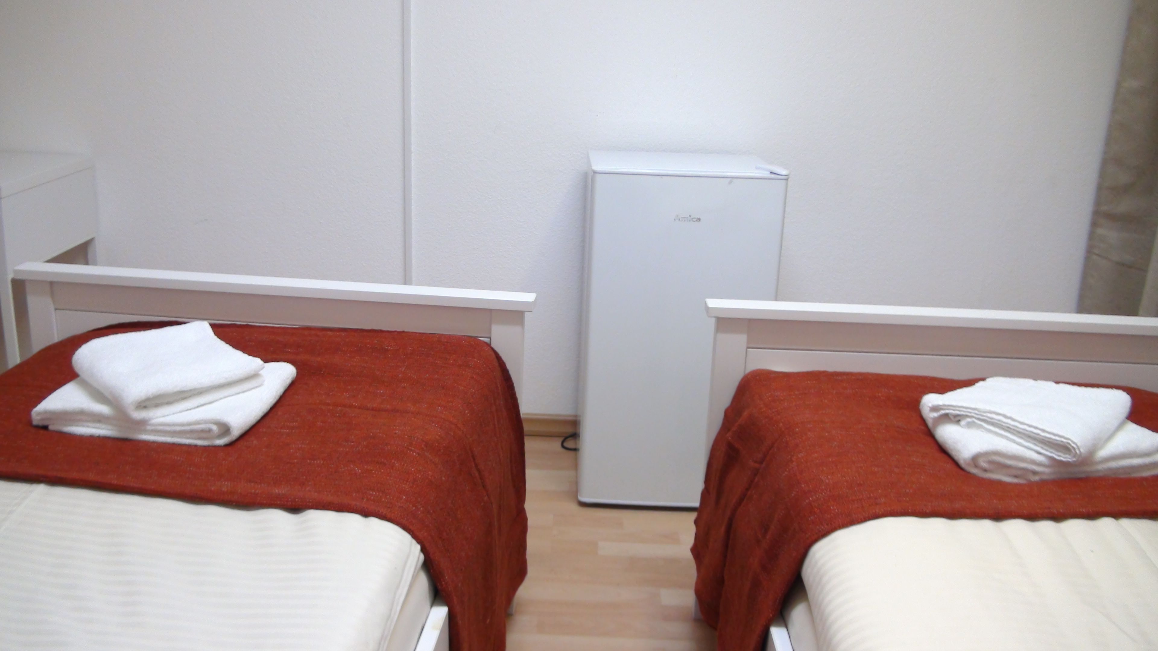 Zwei Einzelbetten mit roter und weißer Bettwäsche, eine weiße Wand und ein weißer Kühlschrank.