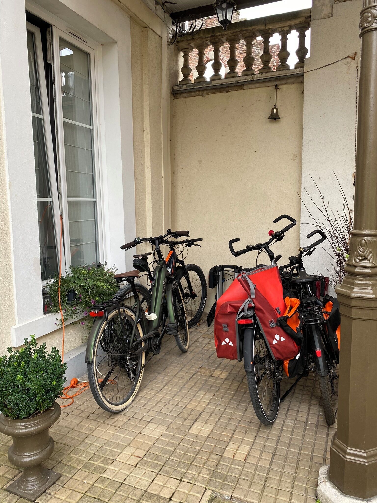 A tiled patio with two bicycles parked against a wall, a potted plant, and a window with curtains.