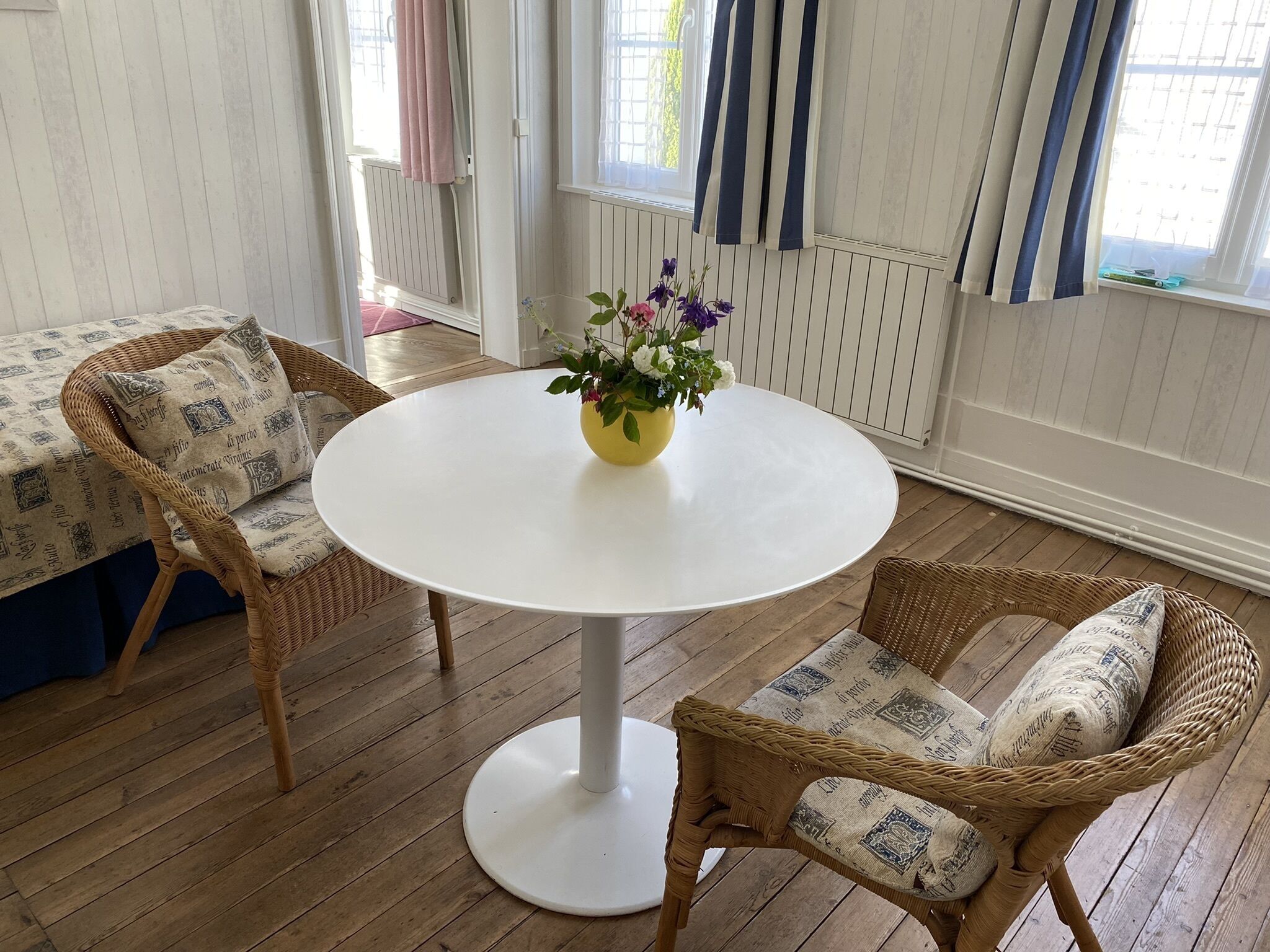 Quarto com mesa redonda branca, duas cadeiras de vime com almofadas estampadas, um vaso com flores e uma janela com cortinas.