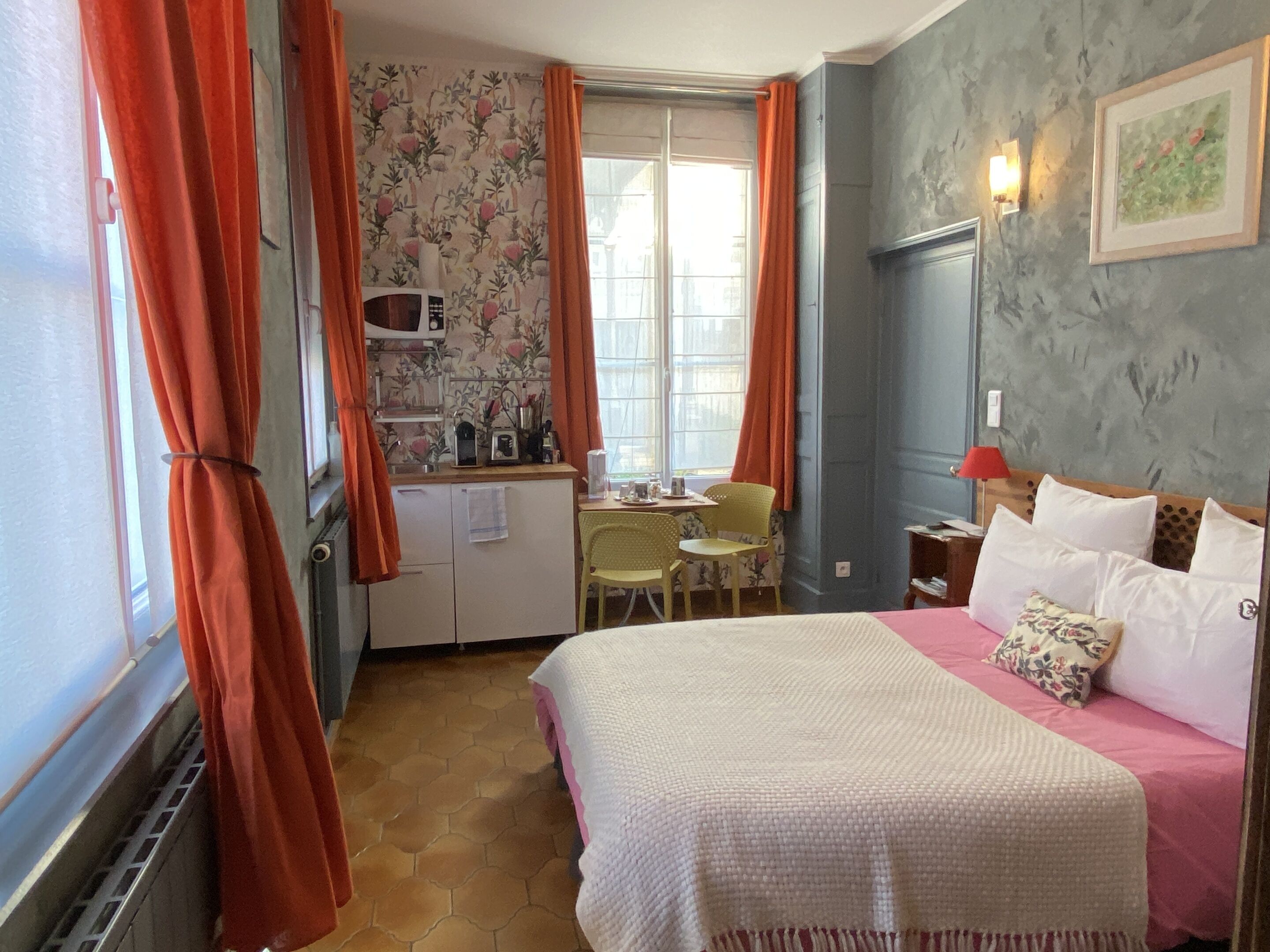Une chambre avec un lit, un bureau et une chaise. Il y a une fenêtre avec des rideaux, un four à micro-ondes et une petite table sur laquelle se trouvent divers objets.