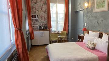 Une chambre avec un lit, un bureau et une chaise. Il y a une fenêtre avec des rideaux, un four à micro-ondes et une petite table sur laquelle se trouvent divers objets.