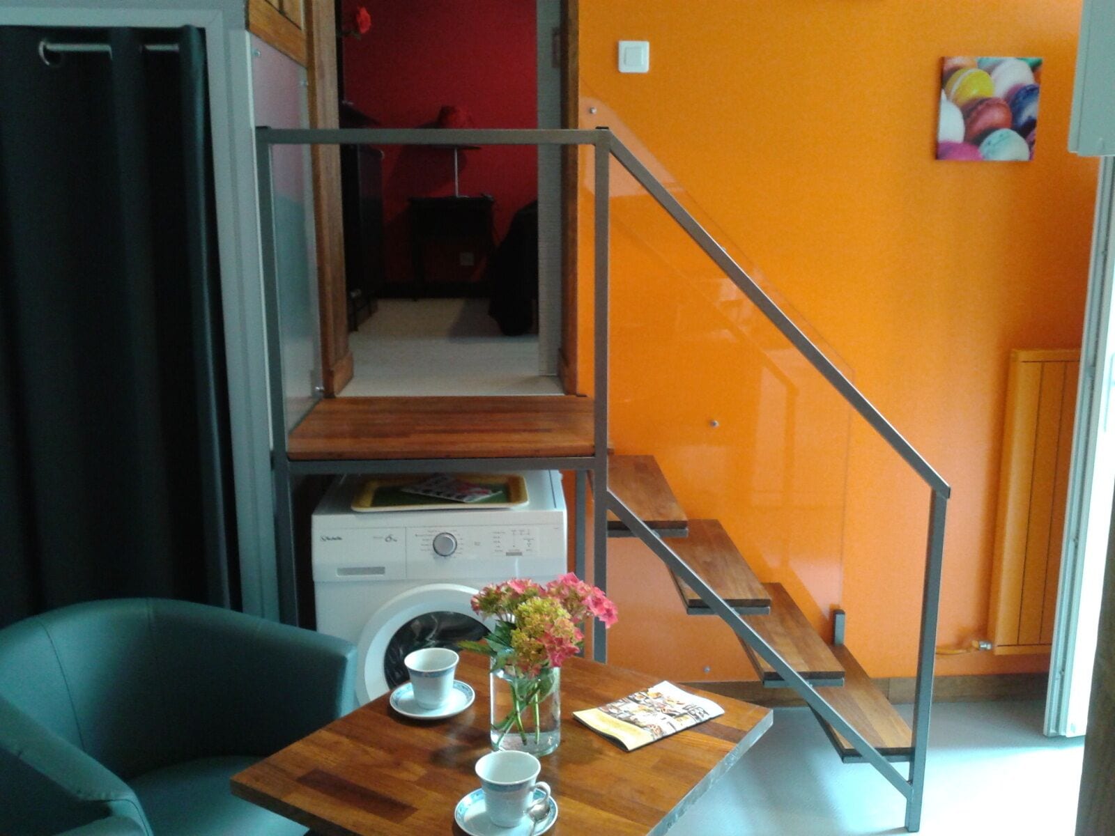 A compact living space with a wooden table, two chairs, a small table with a vase of flowers, and a staircase with a metal railing.