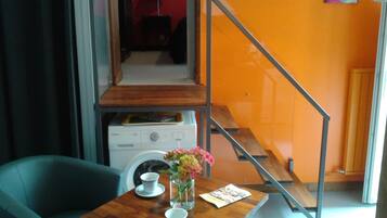 A compact living space with a wooden table, two chairs, a small table with a vase of flowers, and a staircase with a metal railing.
