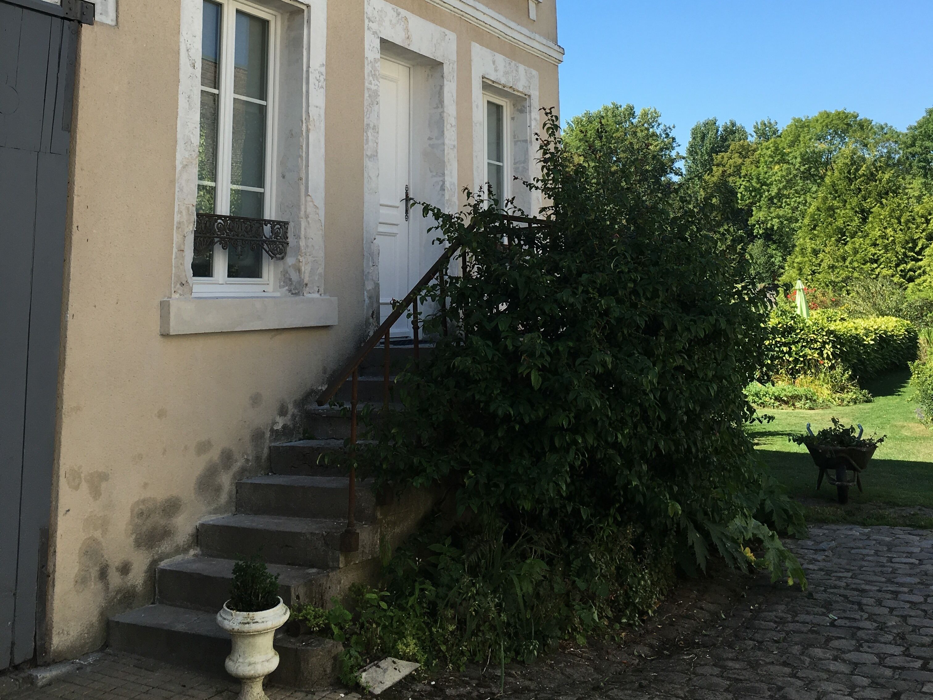 Un edificio con una escalera que conduce a una puerta, rodeado de follaje y un jardín.