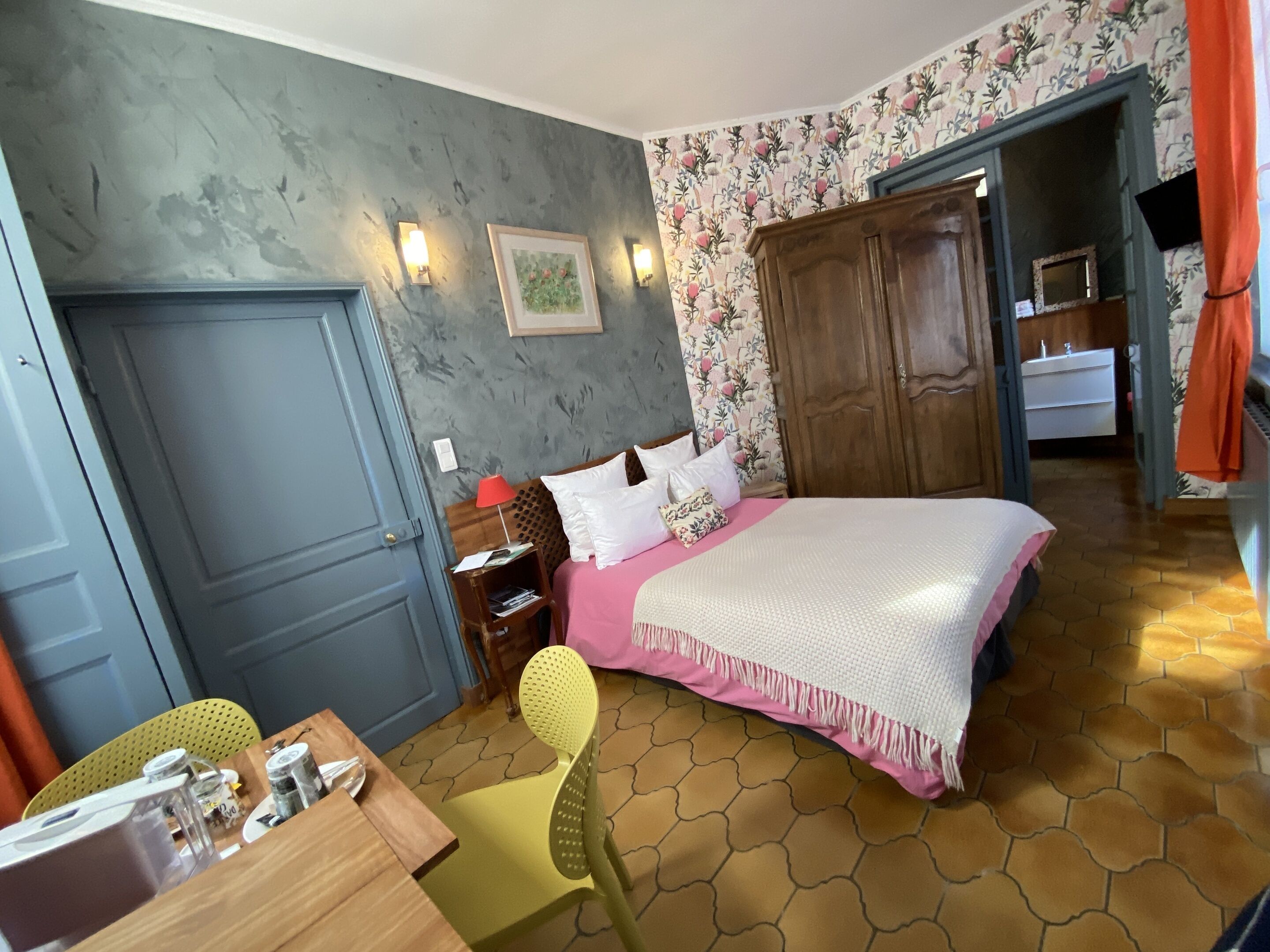 A bedroom with a bed, a nightstand, a chair, a wooden wardrobe, and a bathroom visible through an open door.