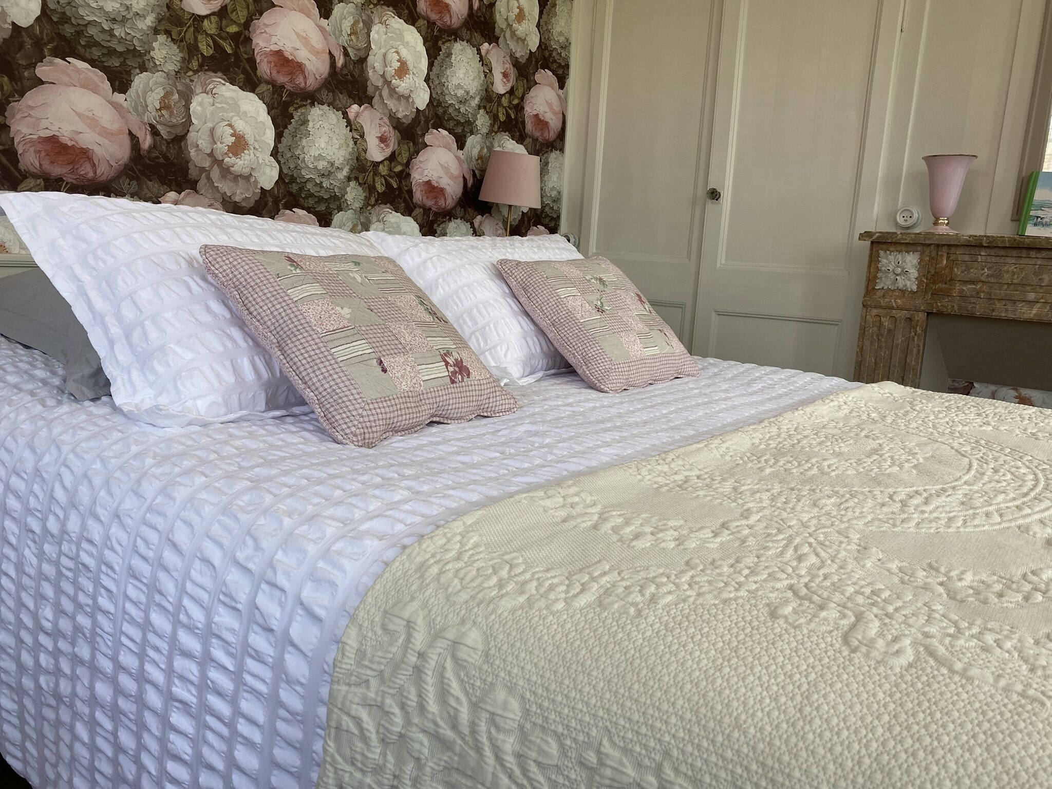 Een netjes opgemaakt bed met een gewatteerde witte dekbedovertrek en gestreepte kussens, een behang met bloemenprint, een bedlamp en een houten nachtkastje met een vaas.