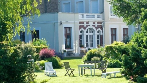 Ein großes, stattliches Gebäude mit einem gepflegten Garten, der eine Rasenfläche, mehrere Stühle und eine Bank zeigt.