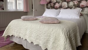 Ein Schlafzimmer mit Bett, Kissen, blumigem Wandpapier und einem Fenster mit Vorhängen.