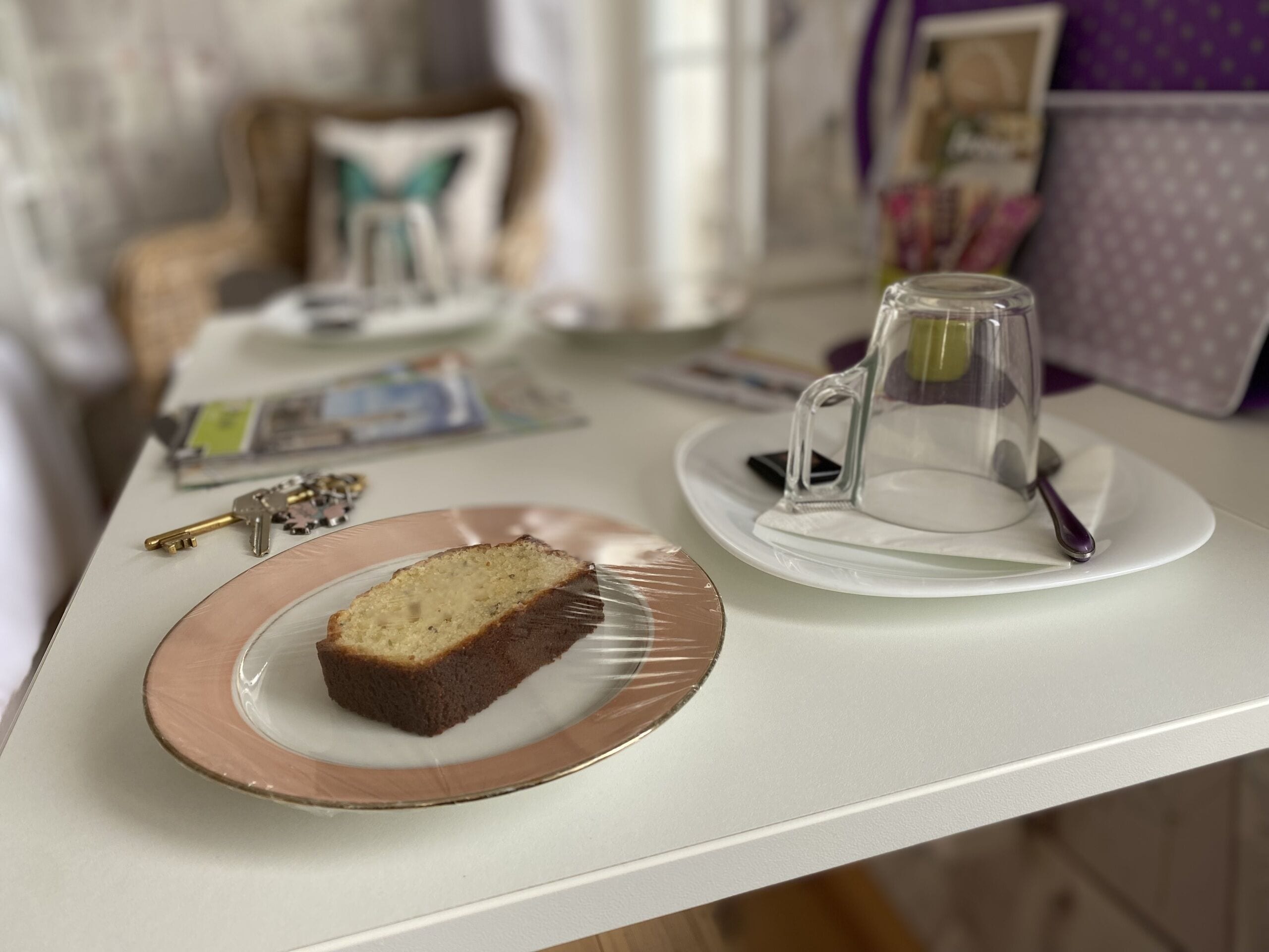 Une part de gâteau sur une assiette, une tasse en verre sur un sous-tasse, des clés et un magazine sur une table.