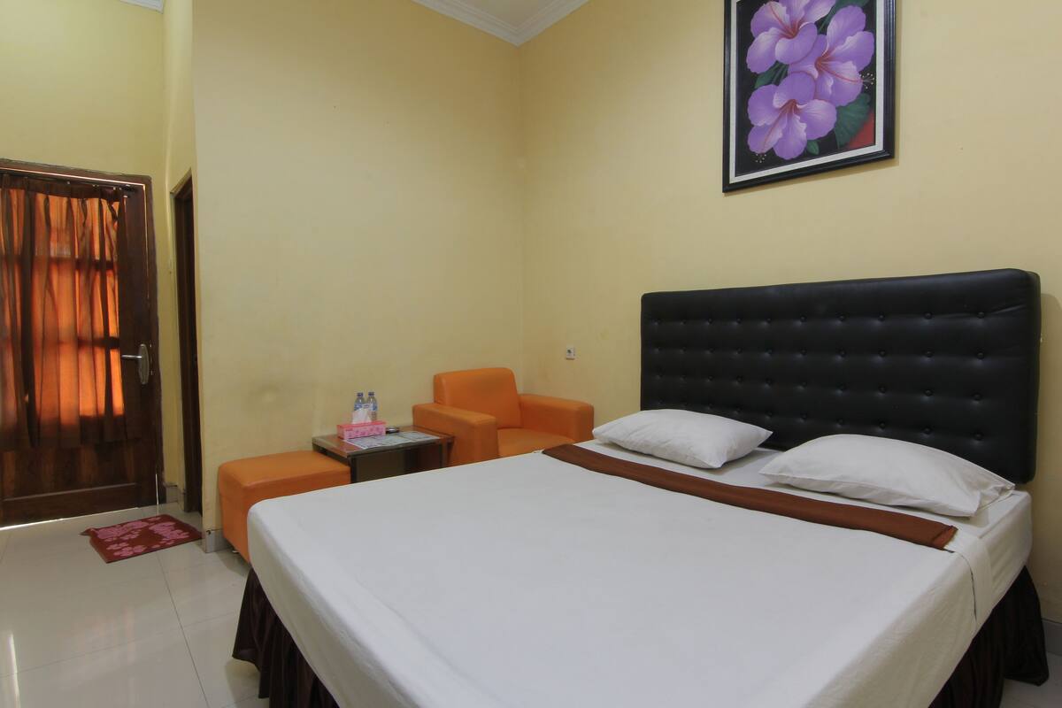 A hotel room with a large bed, an orange armchair, a small table with a bottle and a box, and a door with curtains.