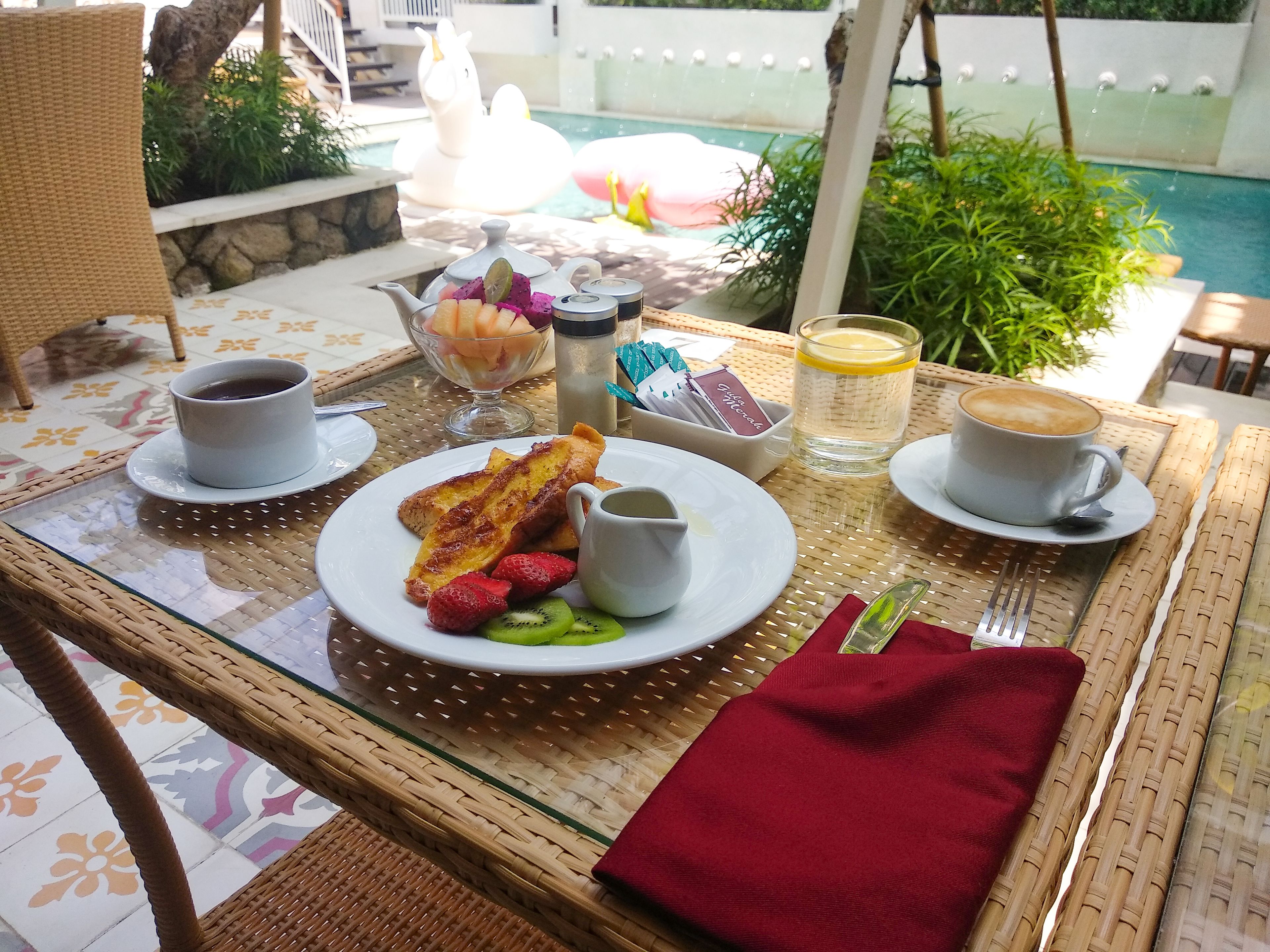 A breakfast setting with a wicker table, a red napkin, a plate with toast, strawberries, kiwi, and a cup of coffee.
