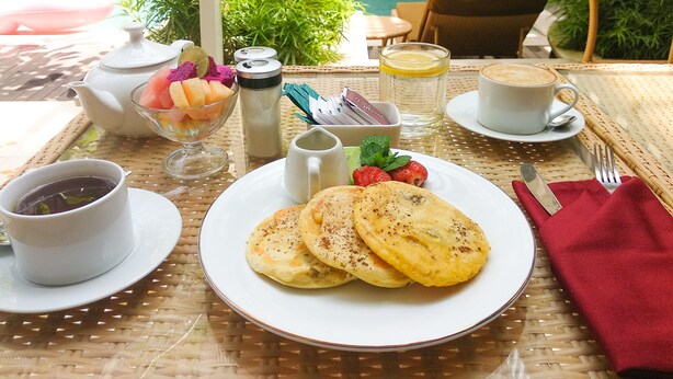 A breakfast setting with a table, a teapot, a cup of coffee, a plate of pancakes, a bowl of fruit, and a glass of water. - Rumah Padi Guest House (Tibubeneng)
