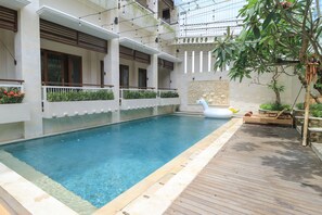 A swimming pool with a wooden deck, surrounded by a white building with balconies and a decorative wall. - Rumah Padi Guest House (Tibubeneng)