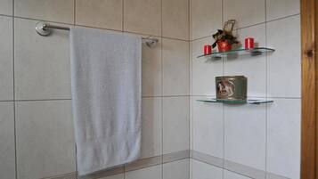 Et badeværelse med et hvidt håndklæde hængende på en metalhåndklæderem, beige flisevægge og to glashylder med dekorative genstande.
