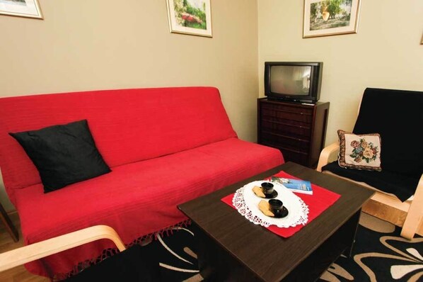 A red sofa with a black pillow, a wooden coffee table with a lace doily and two small cups, a television on a wooden stand, and a chair with a floral pillow. - Vila Izvor (Vrdnik)