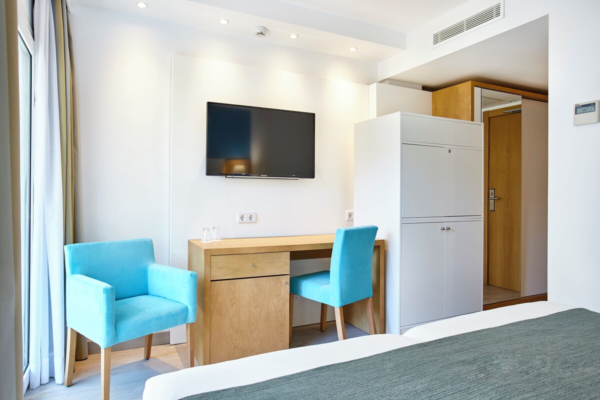 A modern hotel room with a bed, a desk with two chairs, a television, and a wardrobe.