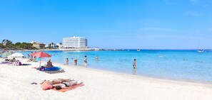 Una playa con gente tomando el sol y nadando, un gran edificio de hotel y barcos en el agua.