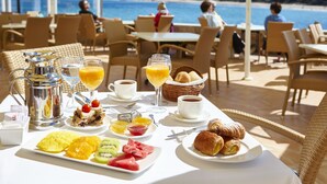 Mesa con alimentos para el desayuno, incluyendo frutas, pasteles y bebidas, con vista a una playa.