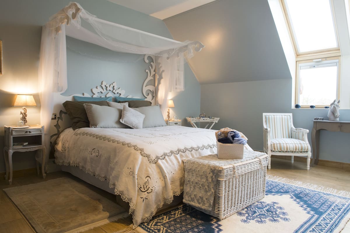 Een slaapkamer met een hemelbed, een rieten nachtkastje, een stoel en uitzicht op de omgeving door een raam.