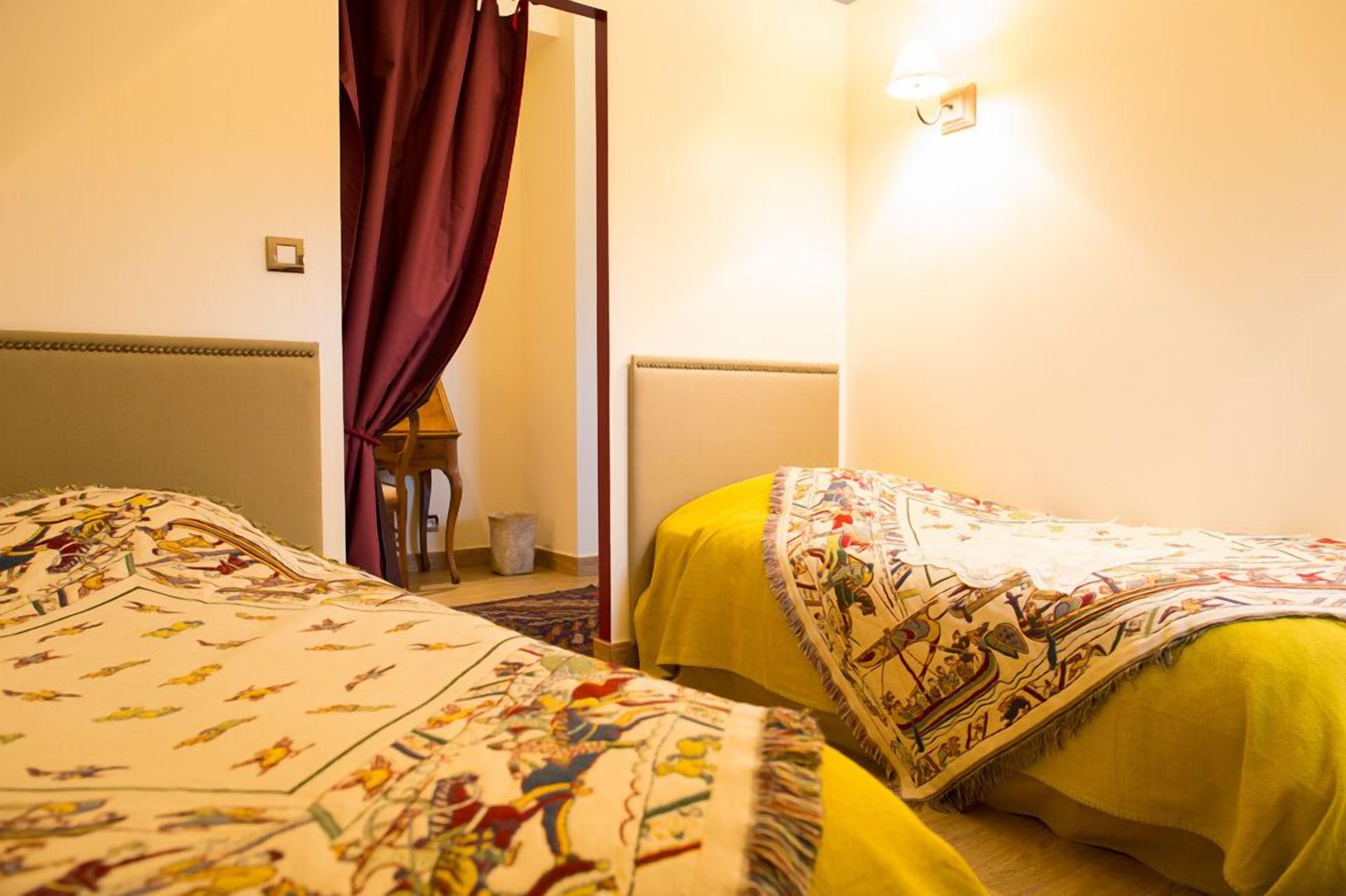 A hotel room with two beds, each with a patterned bedspread, a red curtain, and a wall-mounted light fixture.