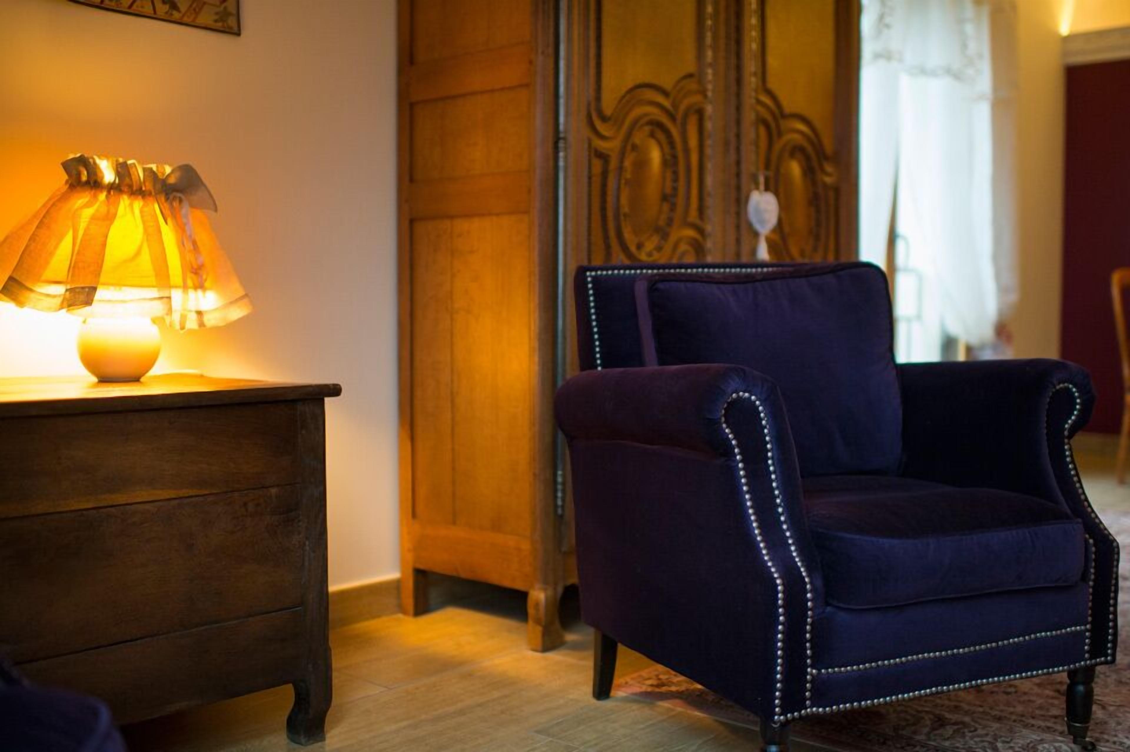A room with a wooden dresser, a dark blue upholstered armchair with decorative stitching, and a lamp with a yellow shade.