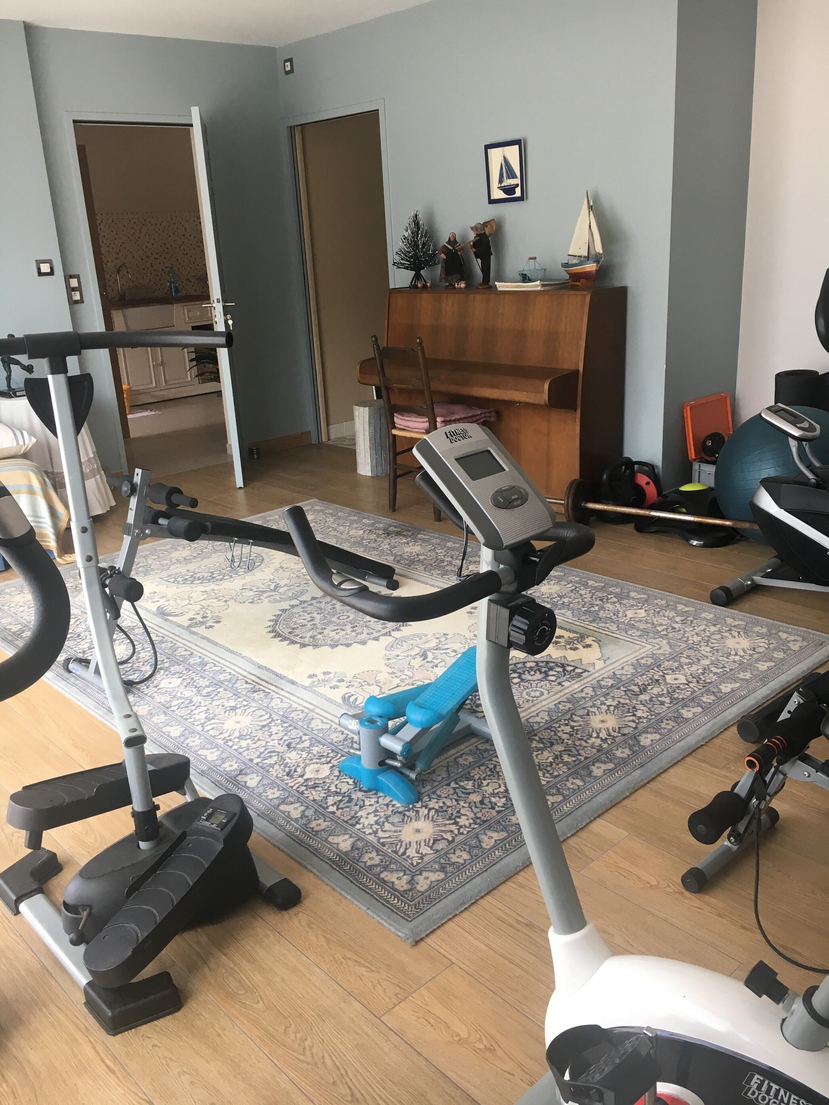 A home gym with an elliptical, a stationary bike, and a treadmill. There is a wooden desk with a chair, a small sailboat decoration, and a patterned rug.