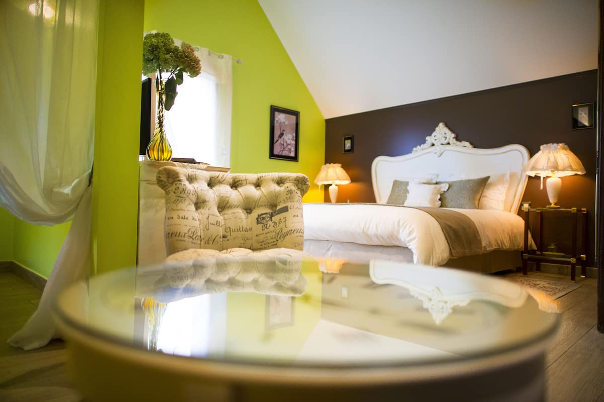 Een slaapkamer met een bed, twee bedlampjes, een nachtkastje, een stoel, een vaas met bloemen, een ingelijste foto aan de muur en een raam met gordijnen.