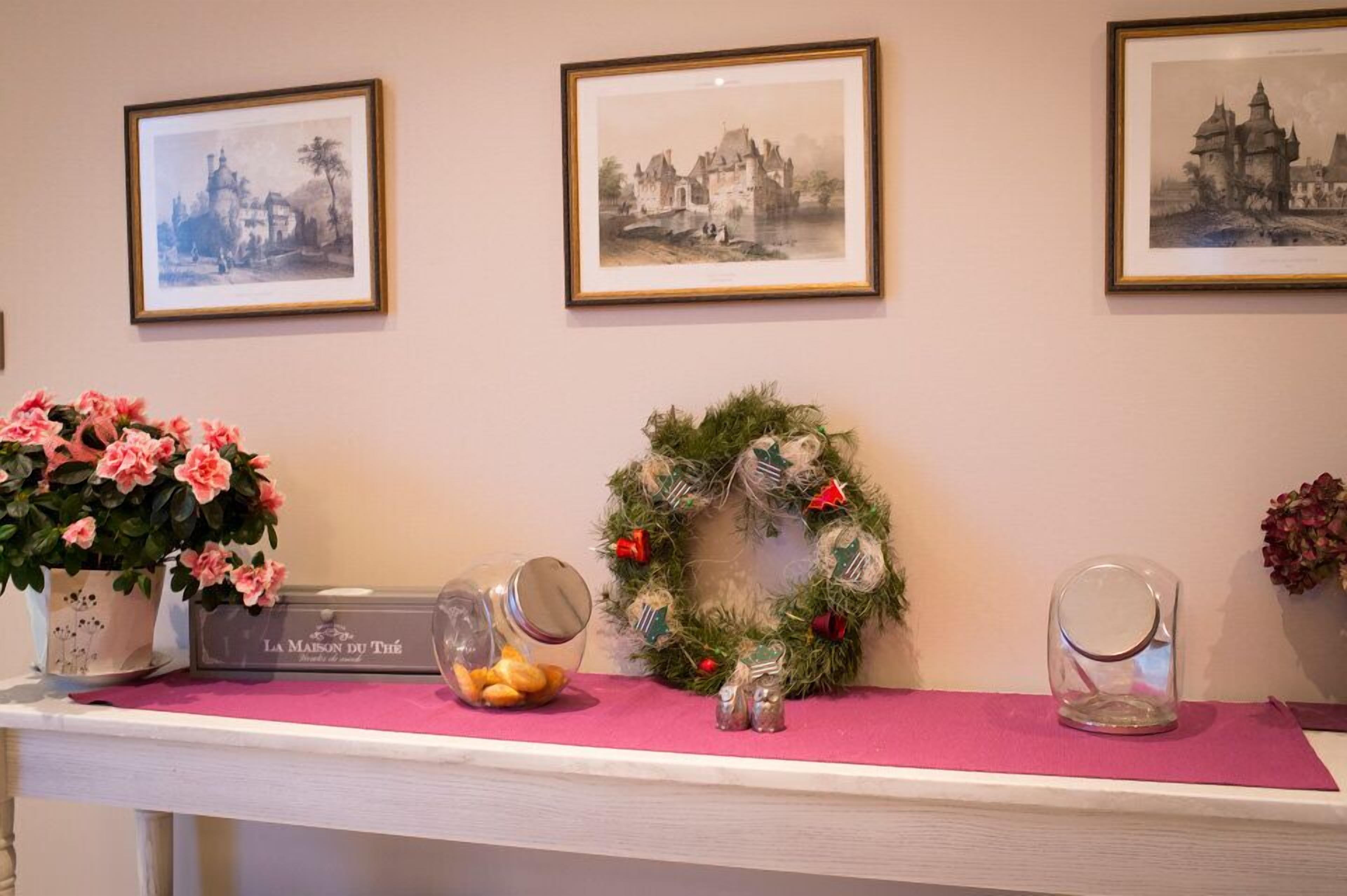 En indrettet reception med indrammede sort-hvide fotografier på væggen, et pink bord med blomsterarrangement, en krans og et glas med nødder.