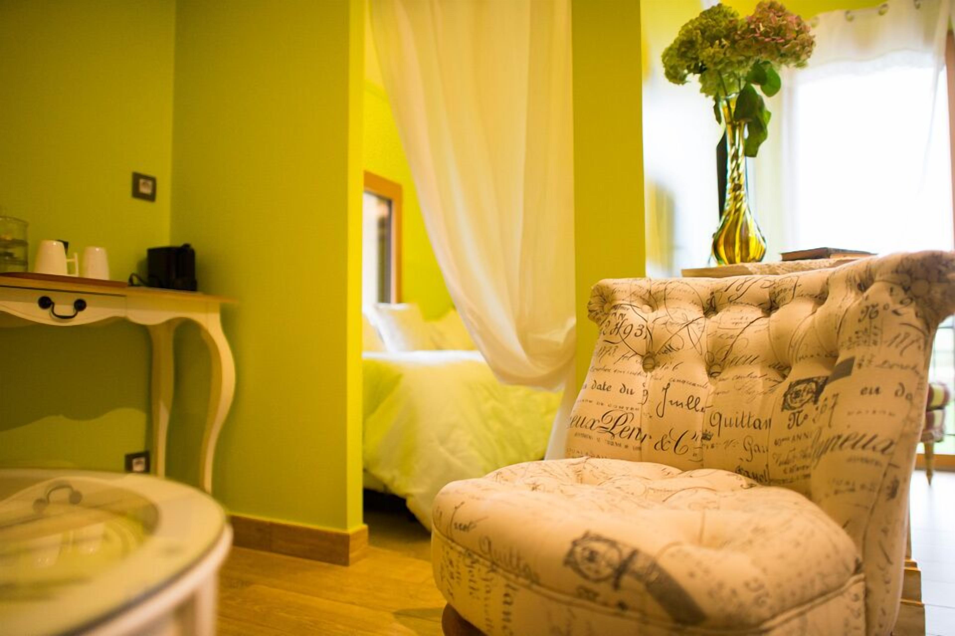 A room with a green wall, a white chair with a pattern, a bedside table, and a bed with white curtains.