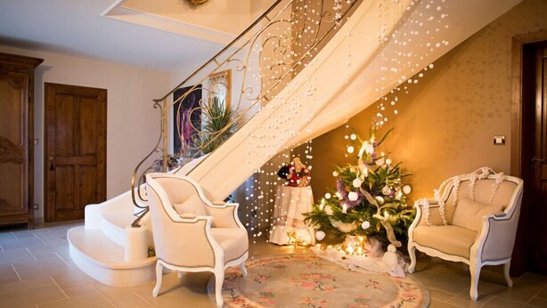Uma escadaria imponente com corrimãos ricamente decorados, uma árvore de Natal e duas poltronas em um ambiente acolhedor, com um tapete floral.