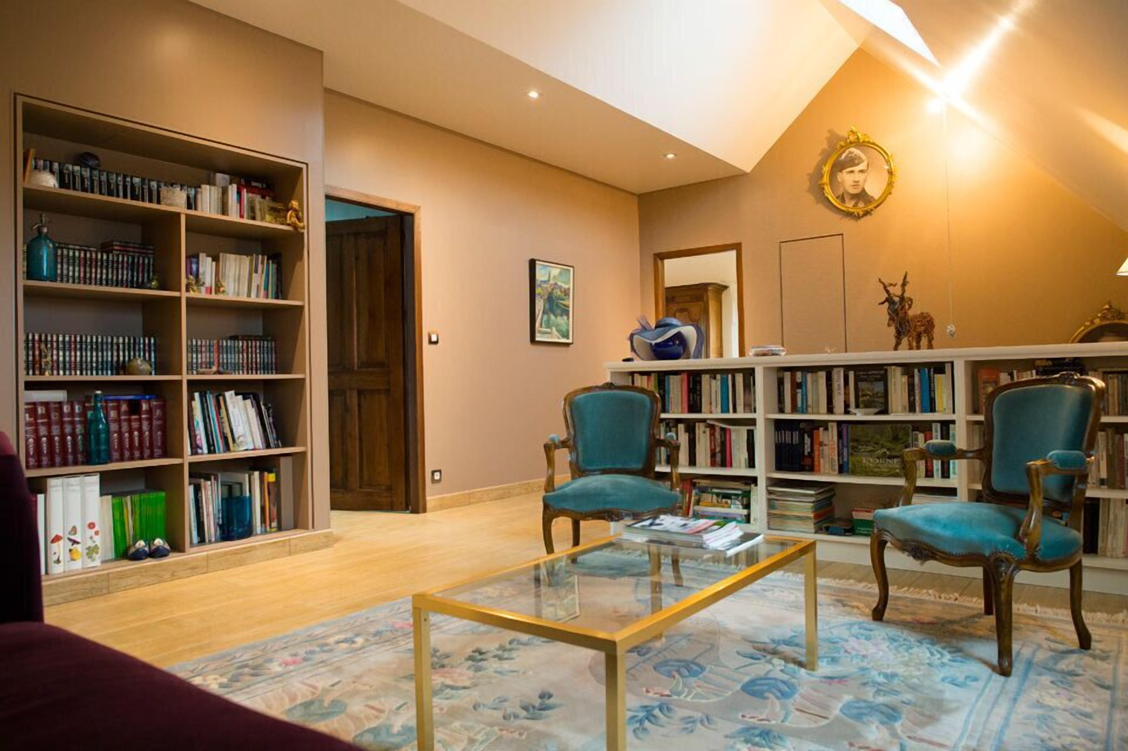 Uma biblioteca com estantes de livros, dois poltronas azuis, uma mesa de centro de vidro e um espelho na parede.