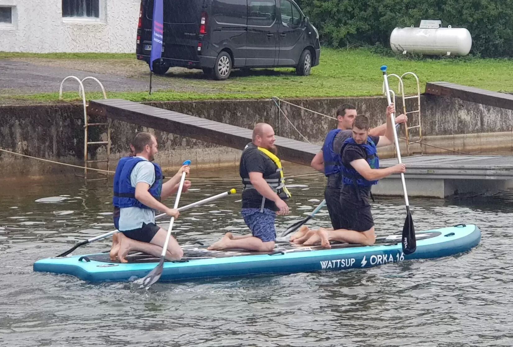 Three people on a paddleboard in a body of water, with a van and a white cylindrical object in the background.