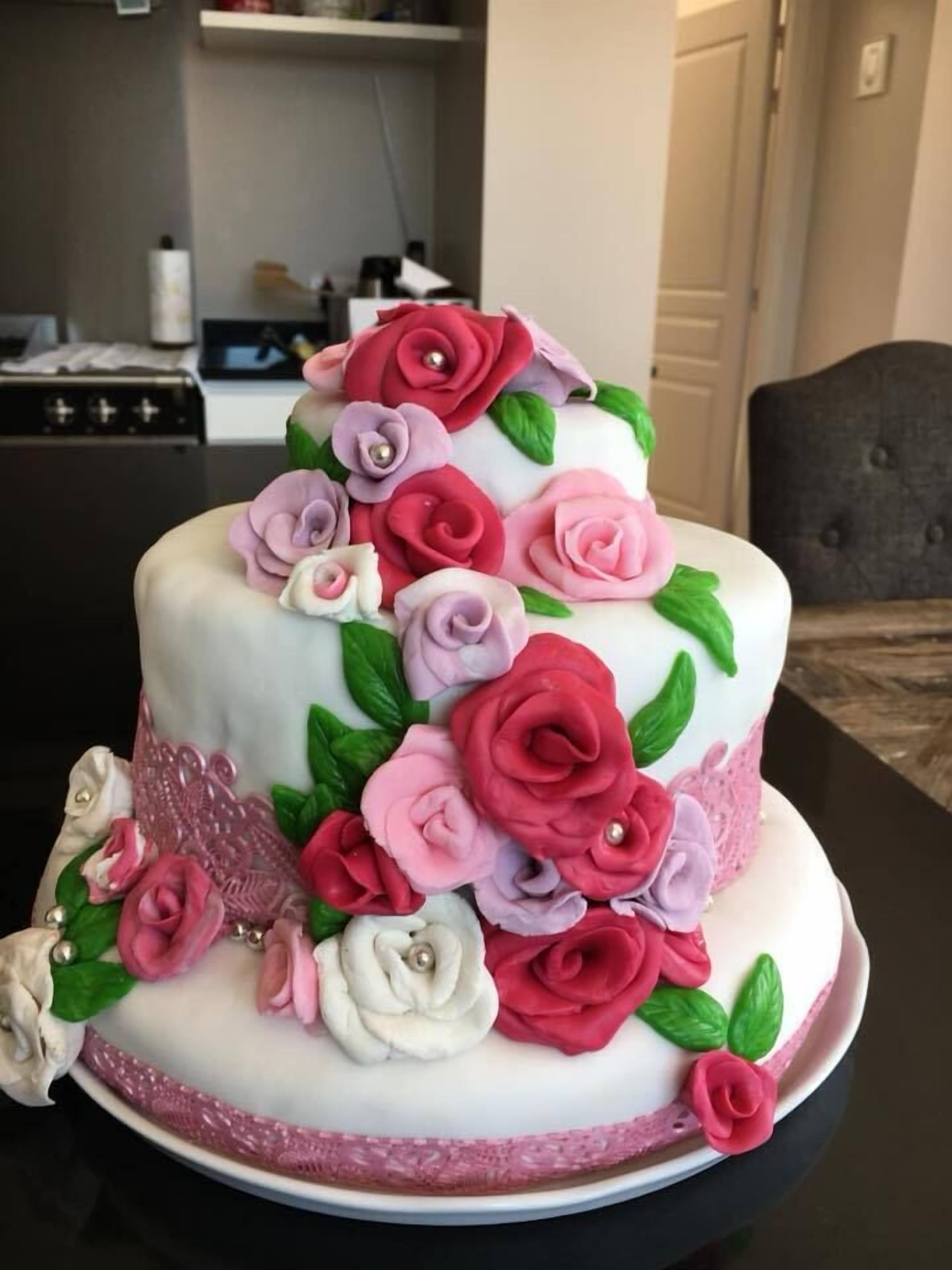 Een tweelaagse taart met roze en rode rozen en groene bladeren op een witte basis, geplaatst op een tafel in een kamer met een keuken op de achtergrond.